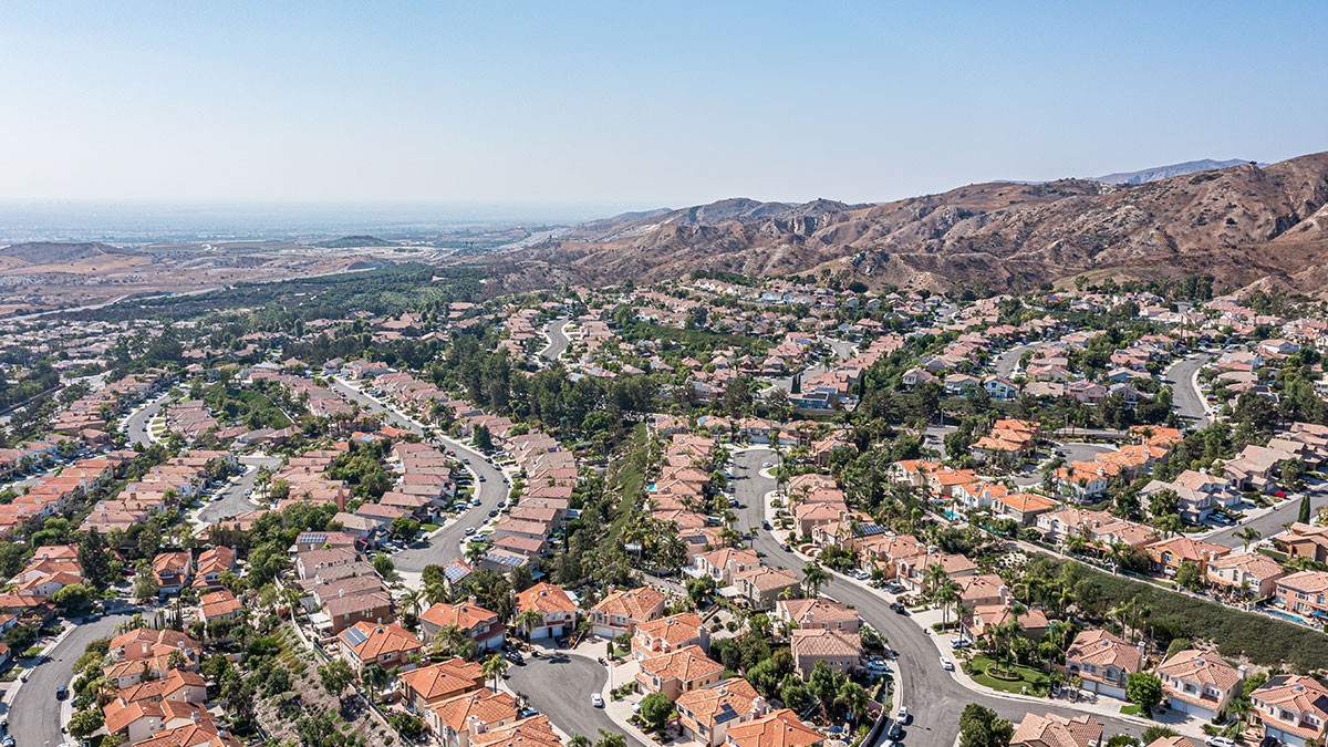 Aerial view of a suburban neighborhood with winding streets, red-roofed houses, and surrounding hills under a clear blue sky.