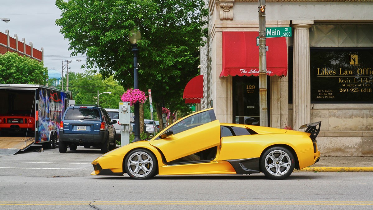 Yellow sports car with scissor doors open, parked on a city street near a law office, under a street sign. Trees and vehicles in the background.