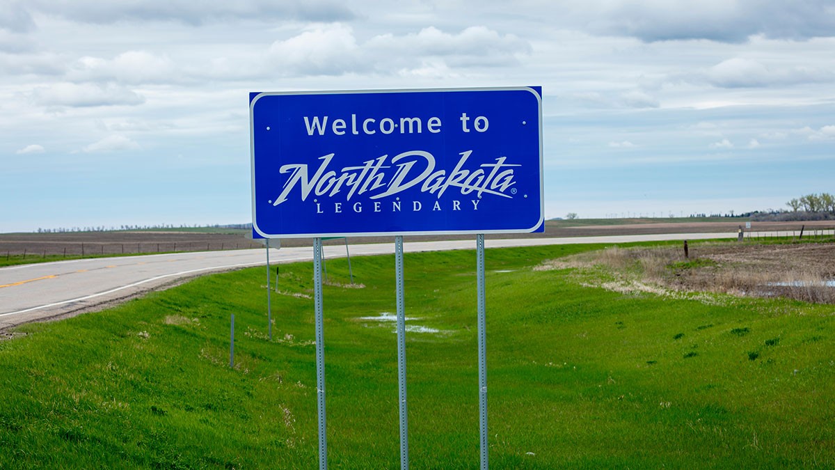 Blue "Welcome to North Dakota Legendary" sign beside an empty rural road, with green grass and cloudy sky in the background.