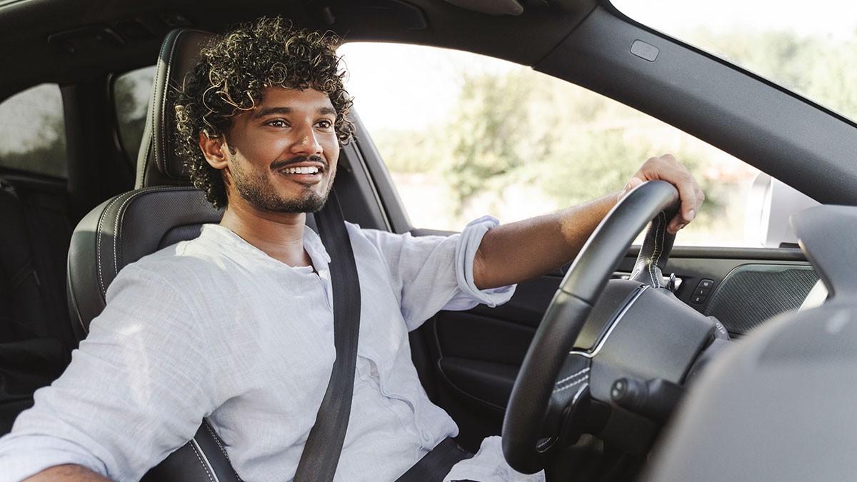 Smiling man with curly hair driving a car, wearing a white shirt and seatbelt.