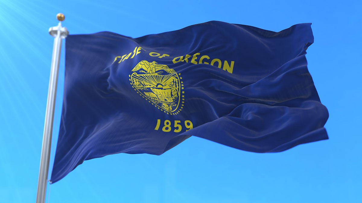 The Oregon state flag waving against a clear blue sky, featuring the state seal and the year 1859 in yellow on a blue background.