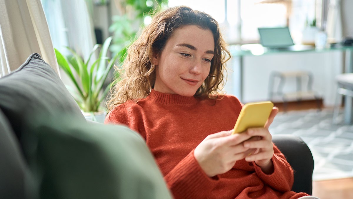 Person in an orange sweater smiling while looking at a yellow phone, sitting on a couch with plants and a desk in the background.