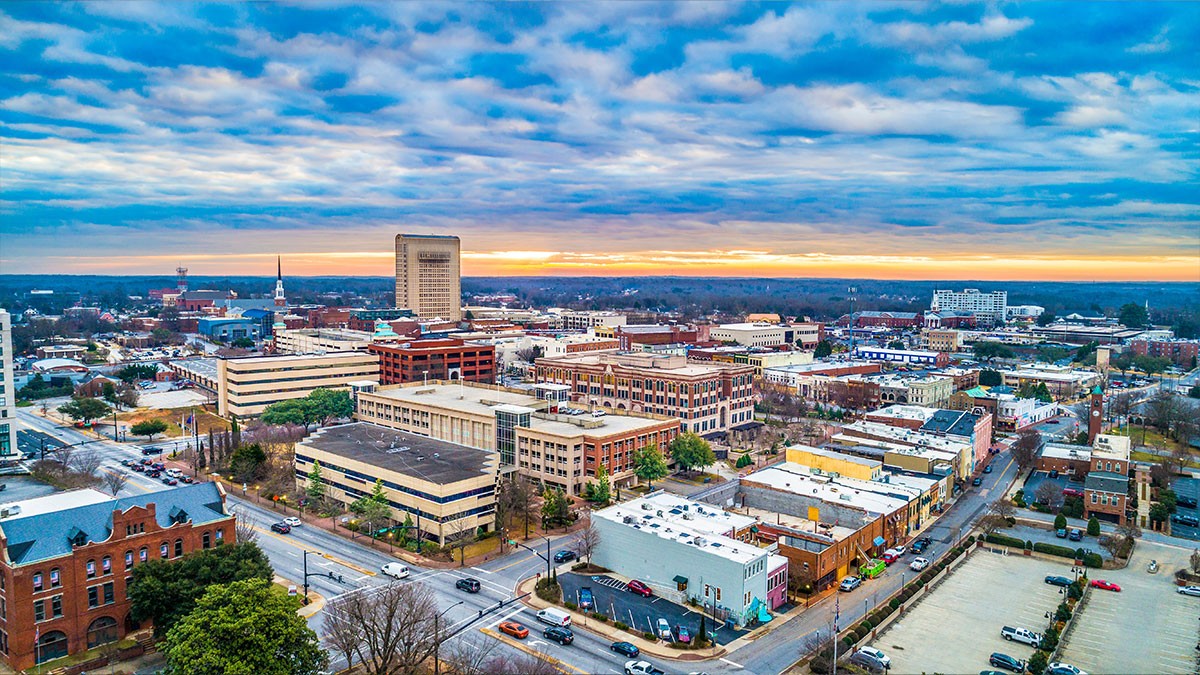 Aerial view of a cityscape at sunset, featuring a mix of modern and historic buildings under a cloudy sky with a horizon glow.