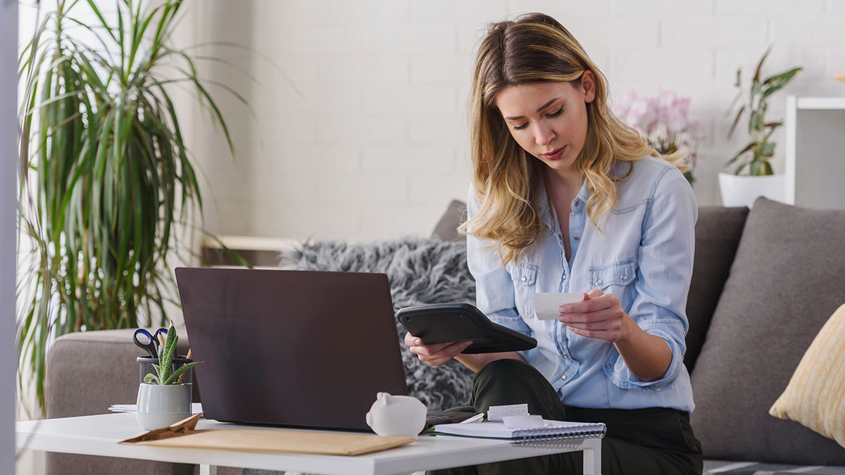Woman in a light blue shirt sits on a couch, holding a tablet and a receipt, with a laptop and notes on a white table in a cozy living room.