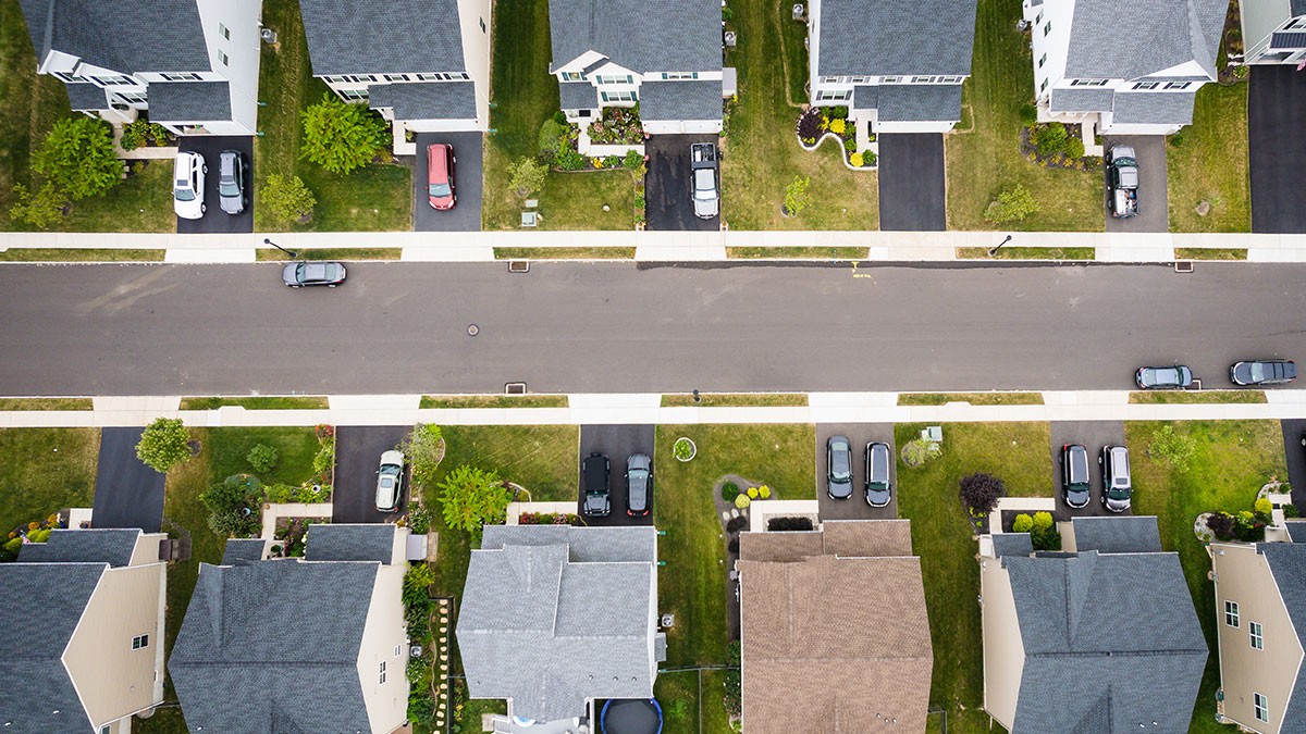 Aerial view of a suburban neighborhood with houses on both sides of a street, driveways, green lawns, and several parked cars.
