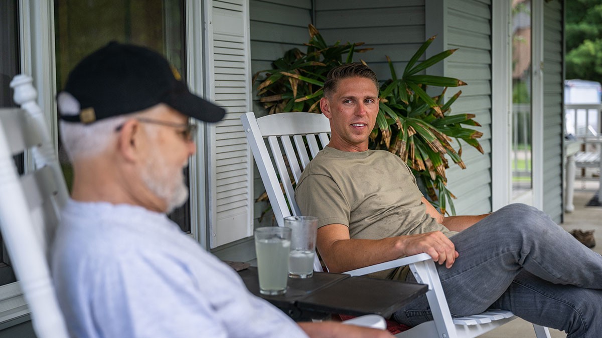 Two men sitting on a porch in rocking chairs, one wearing a cap, engaged in conversation, with a small table holding drinks between them.