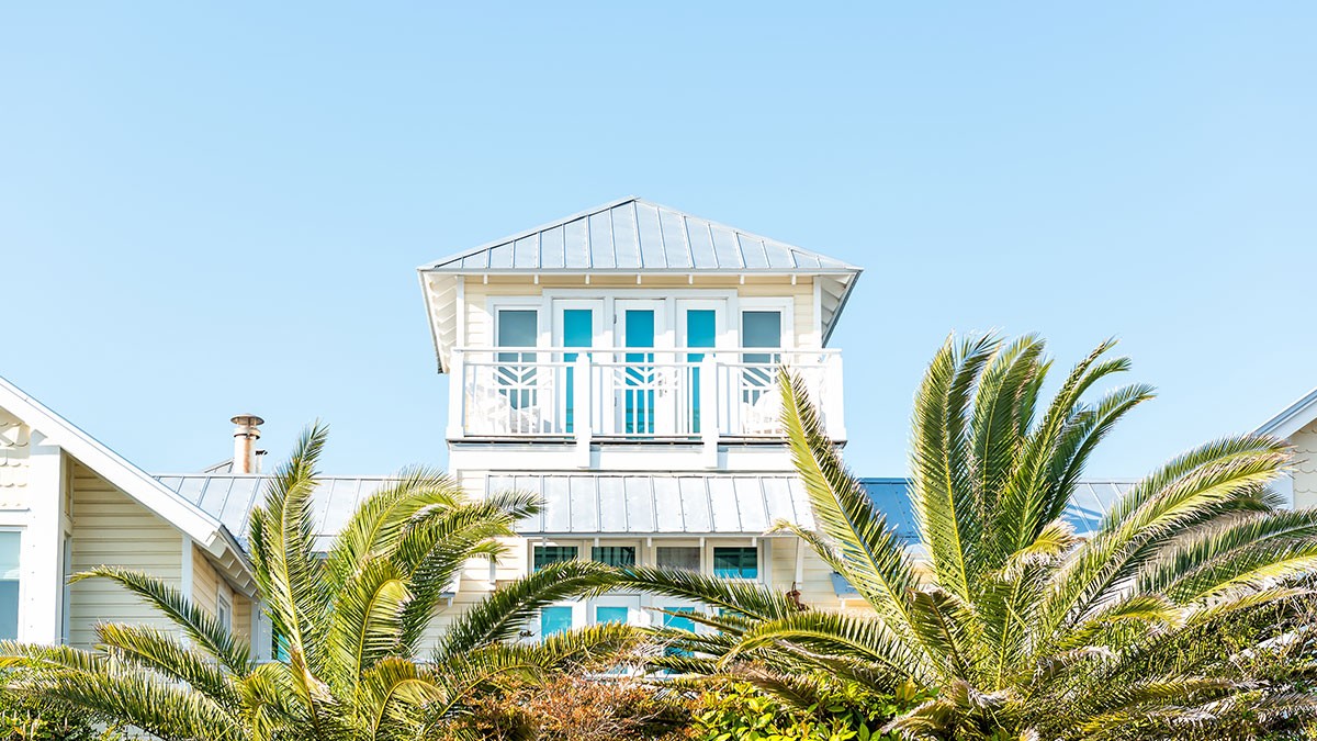 A white beach house with a metal roof and balcony, surrounded by lush palm trees, under a clear blue sky.