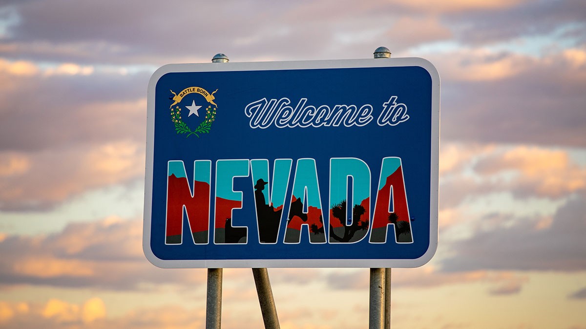A "Welcome to Nevada" sign with silhouettes of desert landscapes, set against a cloudy sky.