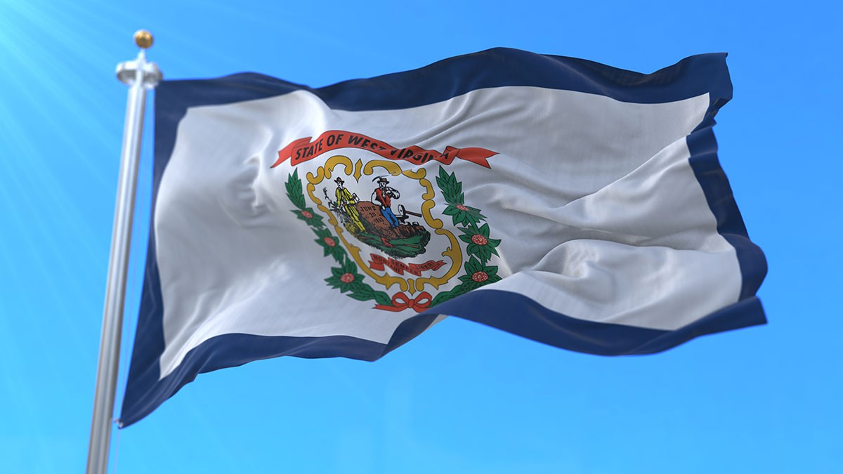 Flag of West Virginia waving against a clear blue sky, featuring the state seal with a miner and farmer surrounded by greenery.