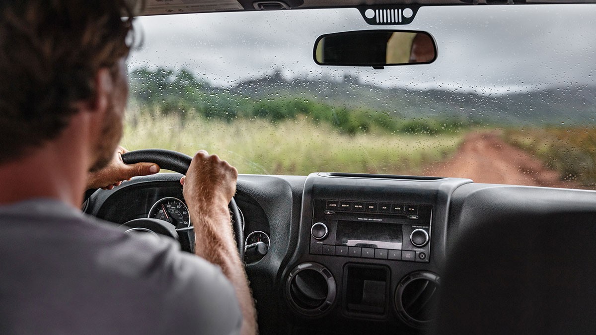 A man drives a car on a muddy road through a lush, rainy landscape, seen from behind. The windshield is speckled with raindrops.