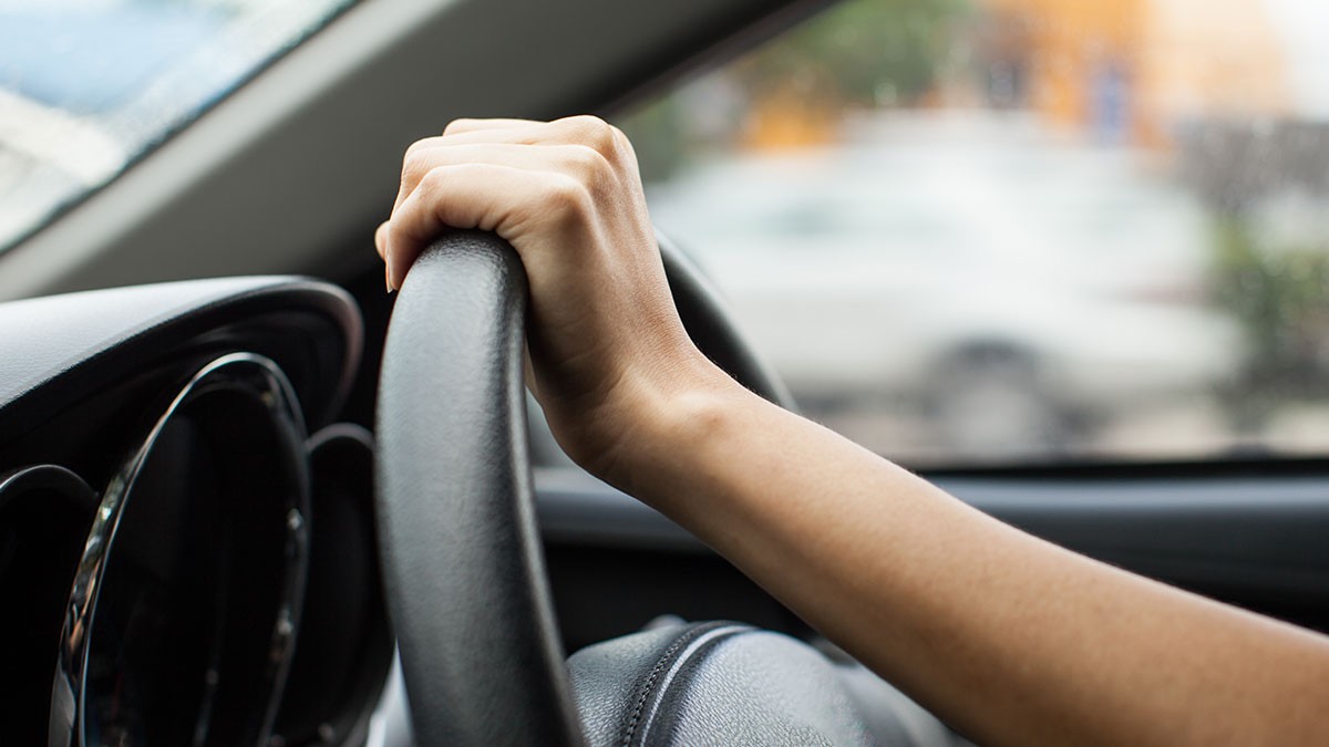Close-up of a person's hand gripping a car steering wheel, with a blurred view of the street visible through the windshield.
