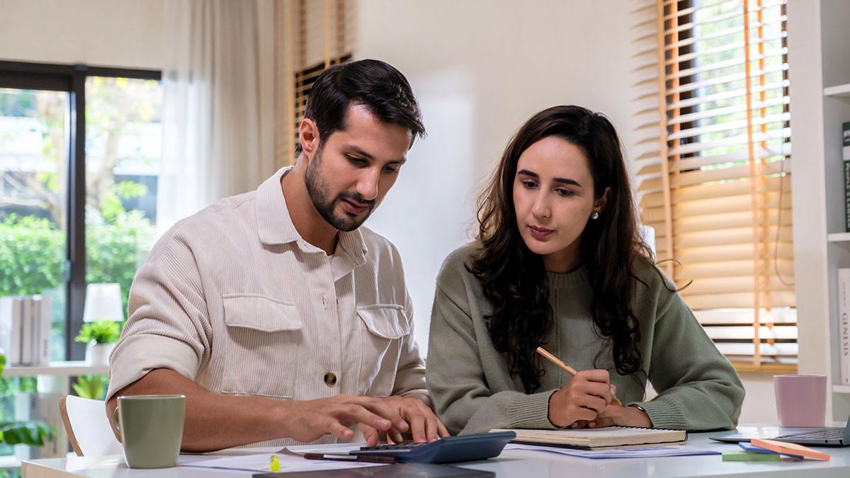 A man and woman sit at a table working on paperwork with a calculator, notebooks, and a mug, in a bright room with open windows.