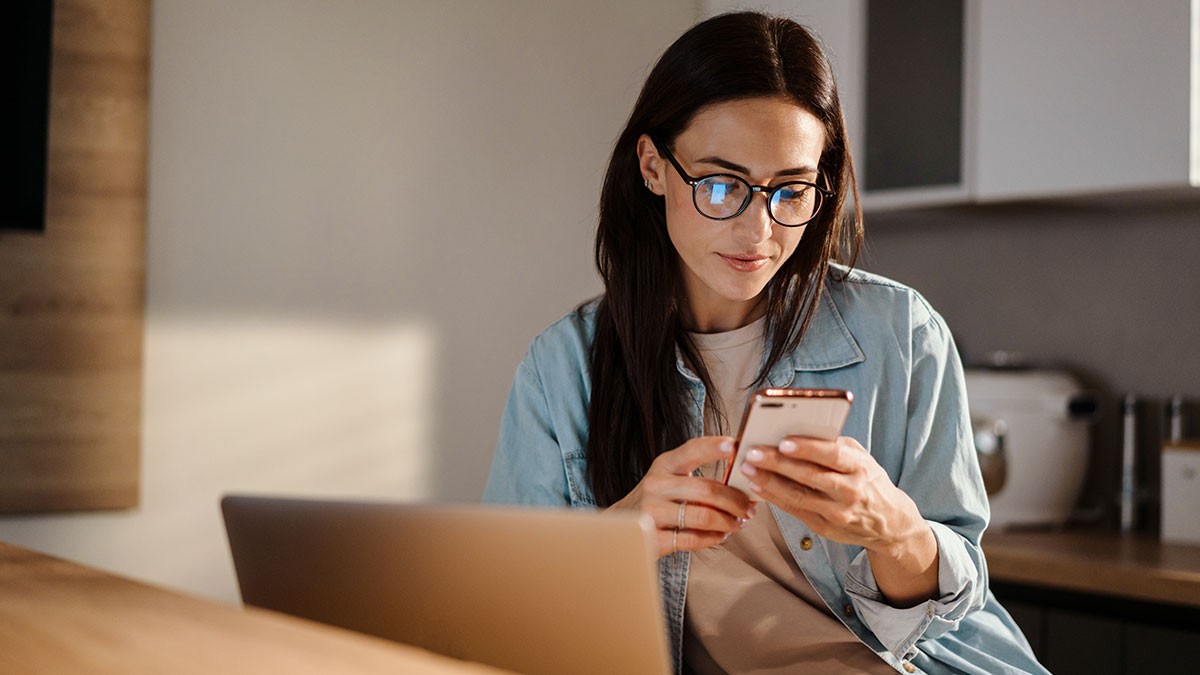 Woman with glasses using a smartphone while sitting next to a laptop in a modern kitchen.