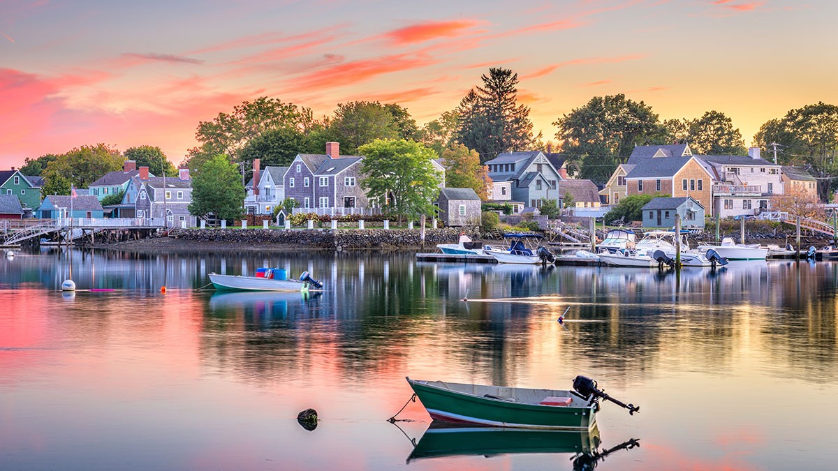 A tranquil harbor at sunset with colorful boats on calm water, and charming houses lining the shore under a pink and orange sky.