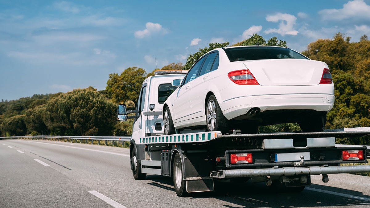 Flatbed tow truck hauling a white sedan on a highway with trees and blue sky in the background.