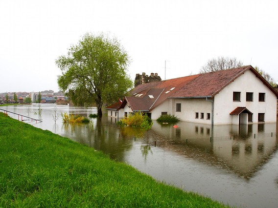 A white house with a red roof partially submerged in floodwaters, surrounded by trees and grass, under an overcast sky.