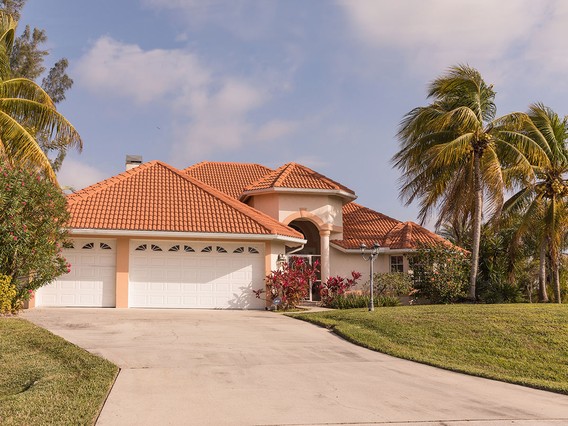 A Mediterranean-style house with a red-tiled roof, palm trees, and a wide driveway leading to a three-car garage.