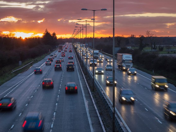 Cars and trucks on a busy highway at sunset, with a vibrant orange sky and streetlights lining the road.