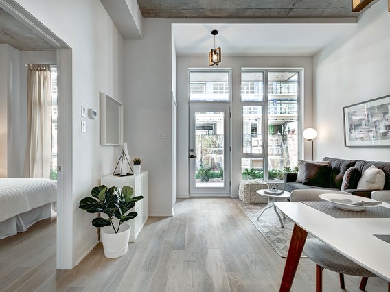 Modern living room with large windows, light wood flooring, and minimalist decor. Partial view of a bedroom and a small indoor plant are visible.