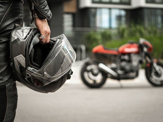 Person in leather jacket holding a motorcycle helmet, with a red motorcycle blurred in the background.
