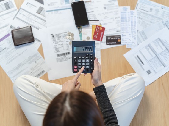 A person sitting on the floor using a calculator with bills, a wallet, a phone, credit cards, and coins scattered around.
