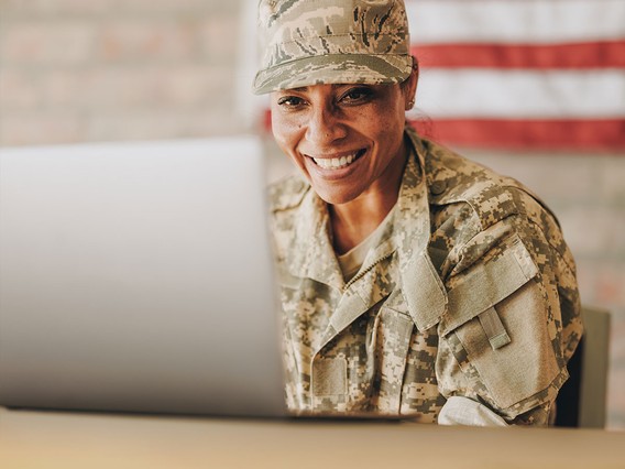 A smiling person in military uniform uses a laptop, with a blurred American flag in the background.