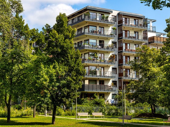 Modern multi-story apartment building with balconies, surrounded by lush green trees and a grassy park area under a blue sky.