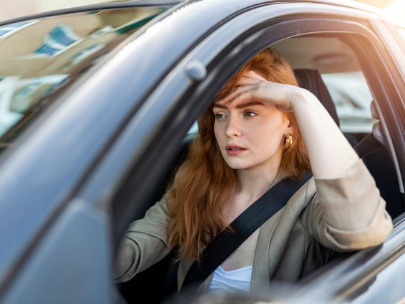A woman with red hair looks thoughtful while sitting in a car, resting her hand on her forehead, wearing a seatbelt and a beige jacket.