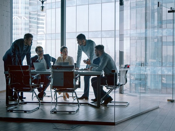 A group of people engaged in a discussion around a conference table in a modern glass-walled office with a city view.