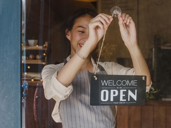 Person smiling while hanging a "Welcome, We Are Open" sign on a glass door, wearing an apron.