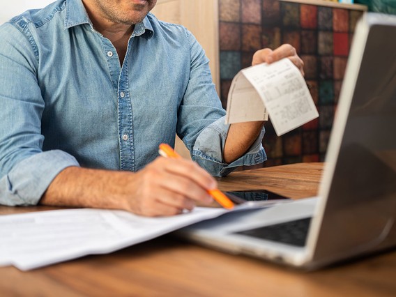Person in denim shirt at desk reviewing a receipt and paperwork, holding a pen beside an open laptop.
