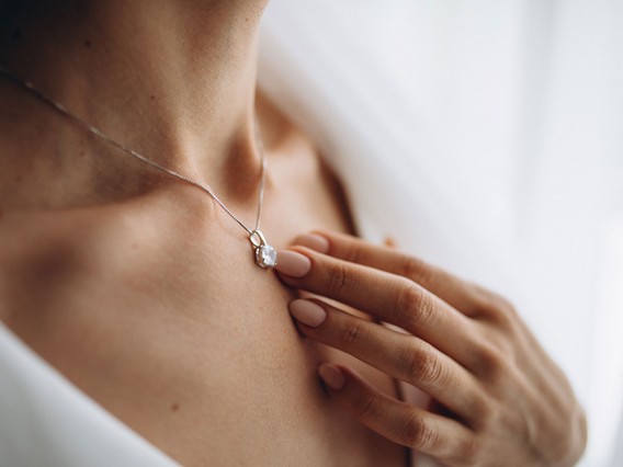 A close-up of a woman wearing an elegant necklace with a pendant, her hand gently touching her chest, showcasing polished nails.