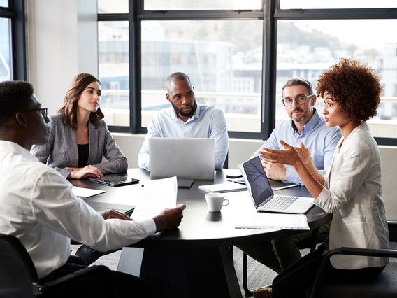 Five colleagues in a meeting around a round table, one woman gesturing as others listen, laptops and papers on the table.