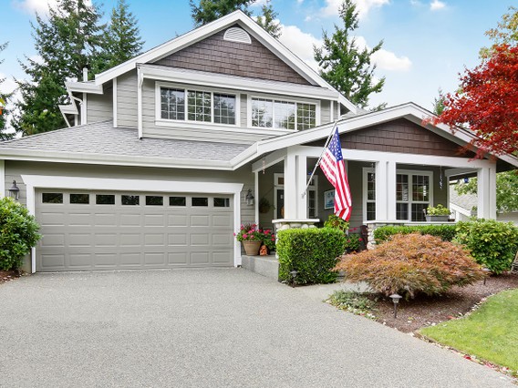 Two-story suburban house with a front porch, American flag, attached garage, and landscaped yard with trees and shrubs.