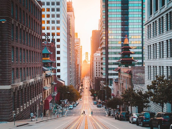 City street at sunset with tall buildings on either side, a distant bridge, and a few people crossing the road.