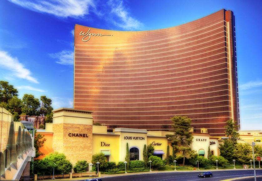 The curved bronze tower of Wynn Las Vegas with the Wynn signature, shops at its base, palm trees and a blue sky