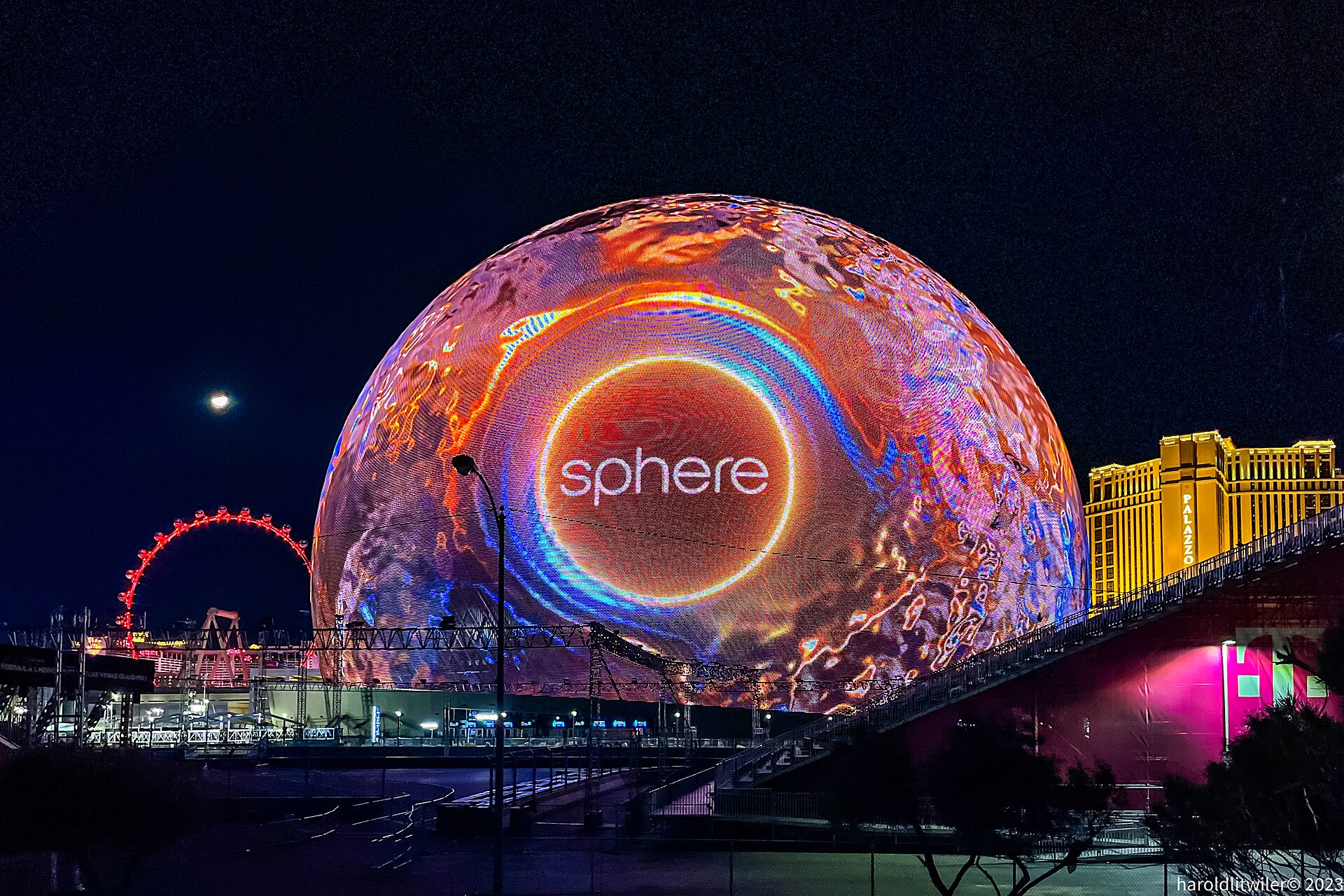 The Sphere glowing orange and blue at night beside the High Roller wheel and the Venetian tower, with race structures still in place