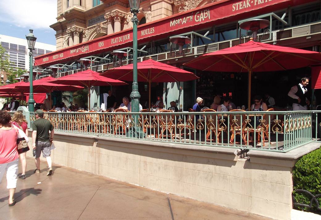 The red awning and red umbrellas of Mon Ami Gabi with diners on the raised patio behind an ornate green railing, beside the Strip sidewalk at Paris Las Vegas