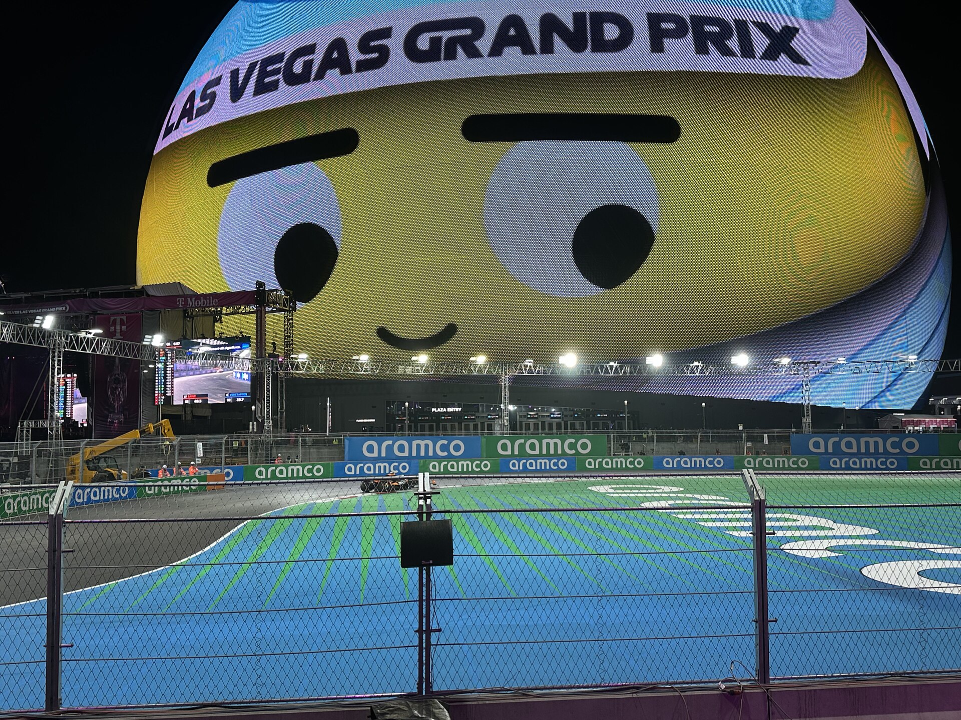 The Sphere lit at night with a cartoon face and the words Las Vegas Grand Prix, with the blue and green run-off and barriers of the track beside it, a crane and fences