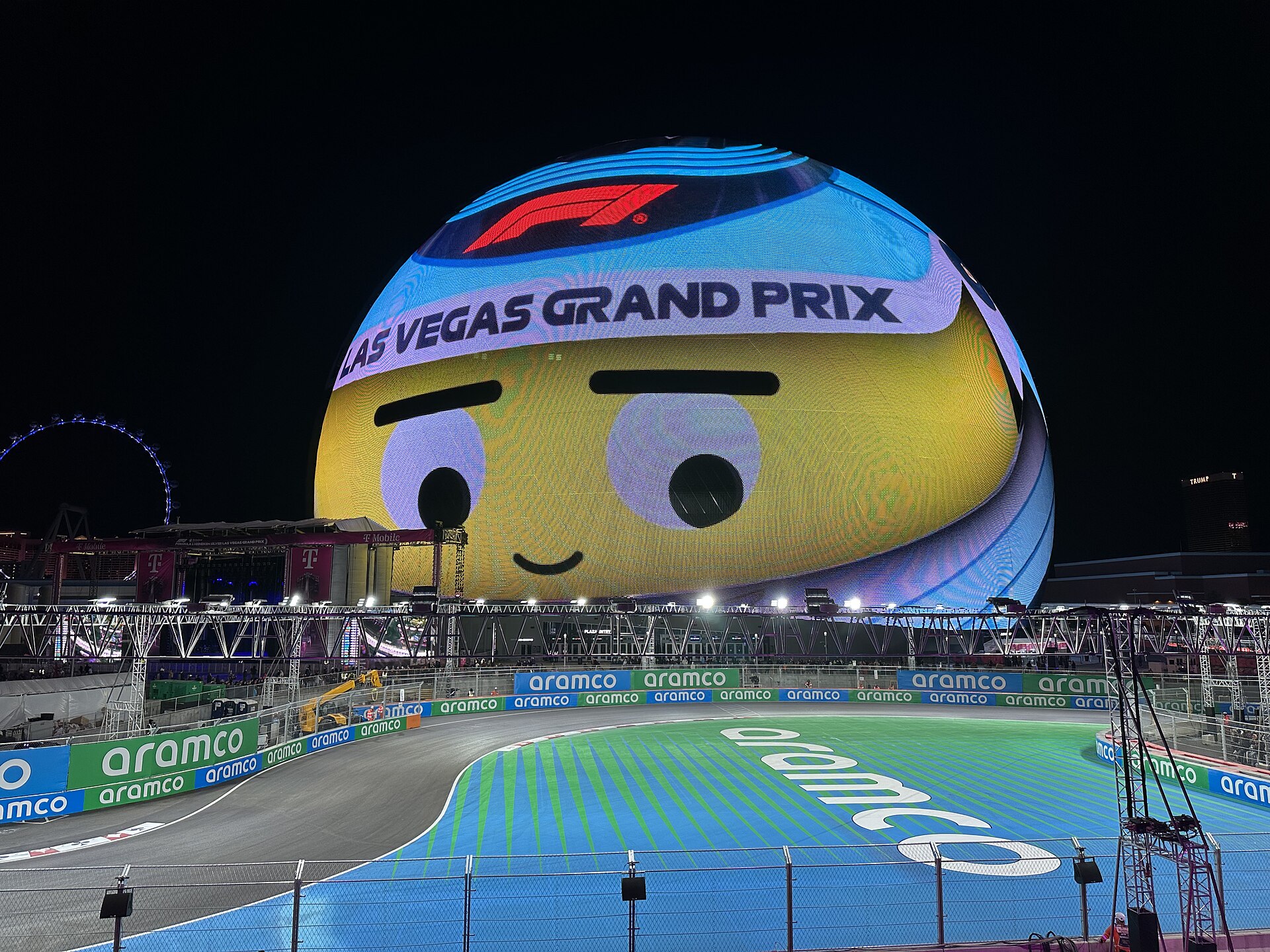 The Sphere at night on race day, with the F1 logo and Las Vegas Grand Prix above a cartoon face, the curving track in front and the High Roller wheel lit on the left