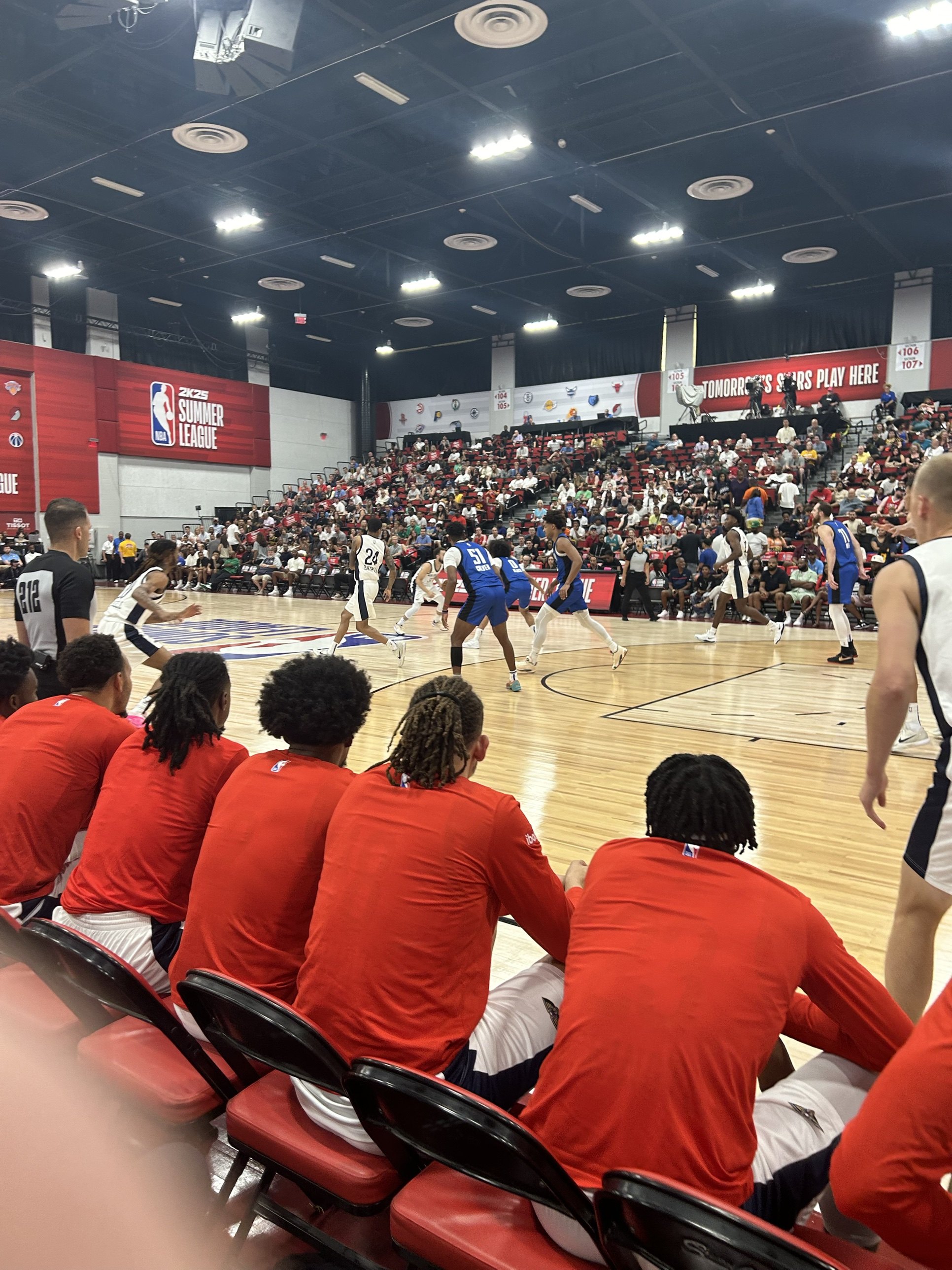 A Summer League game seen from just behind a team bench, players in red warm-up shirts in the foreground and a crowded small arena beyond