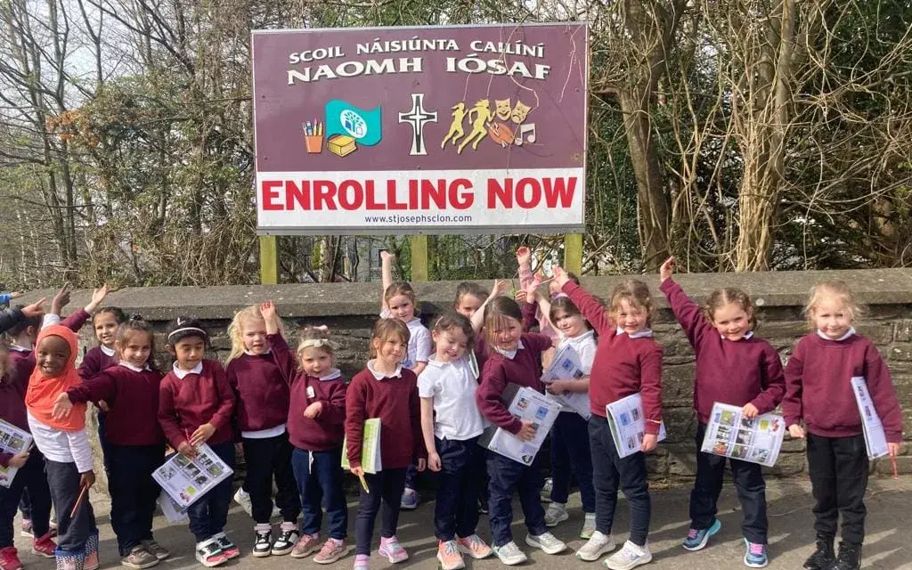 Junior pupils waving beside the school's Enrolling Now sign