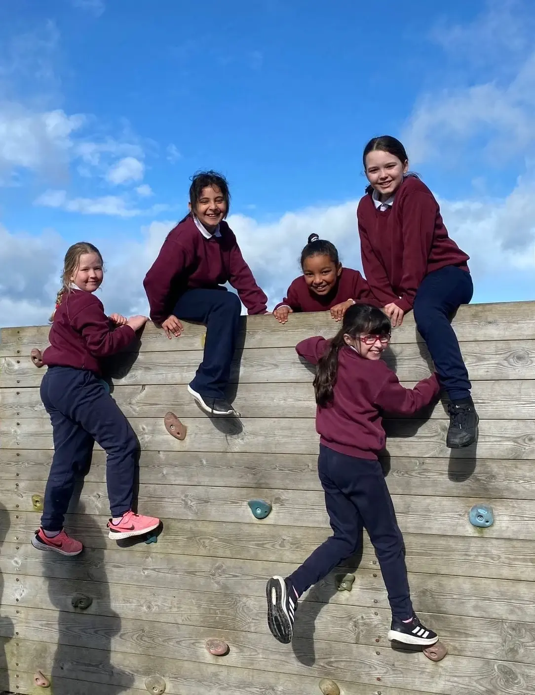 Five smiling pupils in school uniform climbing a wooden climbing wall on a sunny day
