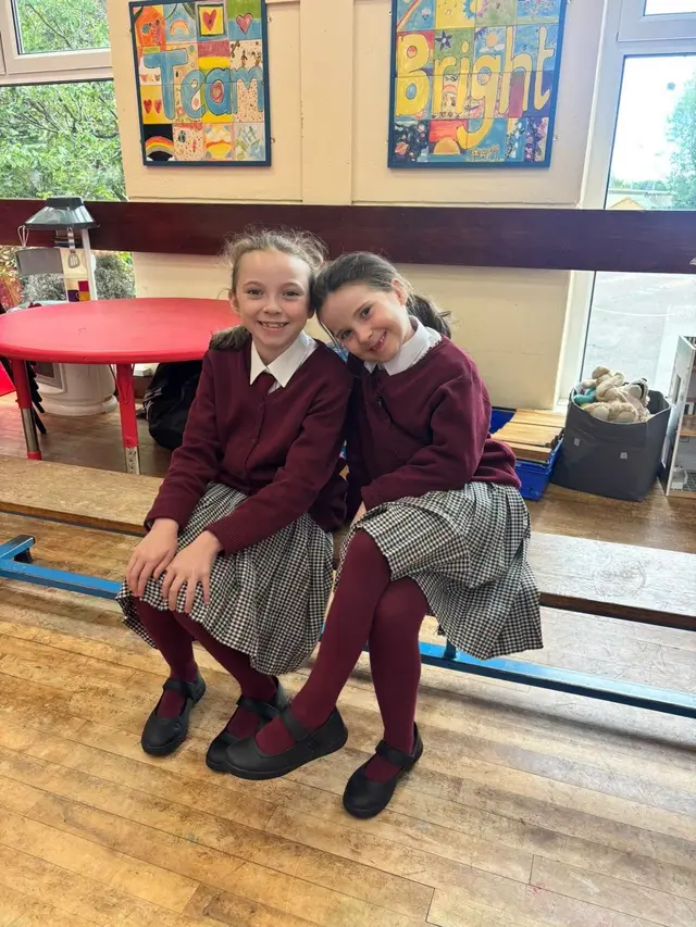 Two pupils in the school uniform: wine cardigan, check pinafore, white shirt, tie and black shoes
