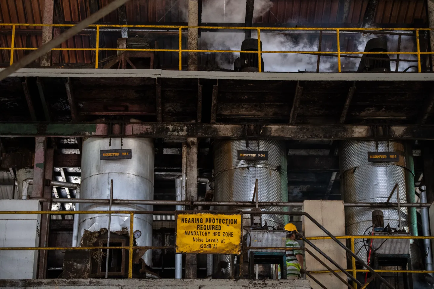 Storage tanks and yellow railings inside a palm oil refinery, with a hearing protection sign