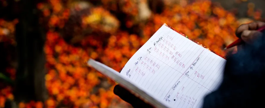 A hand holding a notebook of handwritten figures over a pile of harvested oil palm fruit