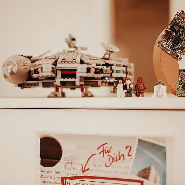 Auf einem Schrank steht ein Playmobil Schiff von Star Wars sowie ein aus Pappe gefertigtes C. davor stehen einige Playmobilfiguren von Star Wars.