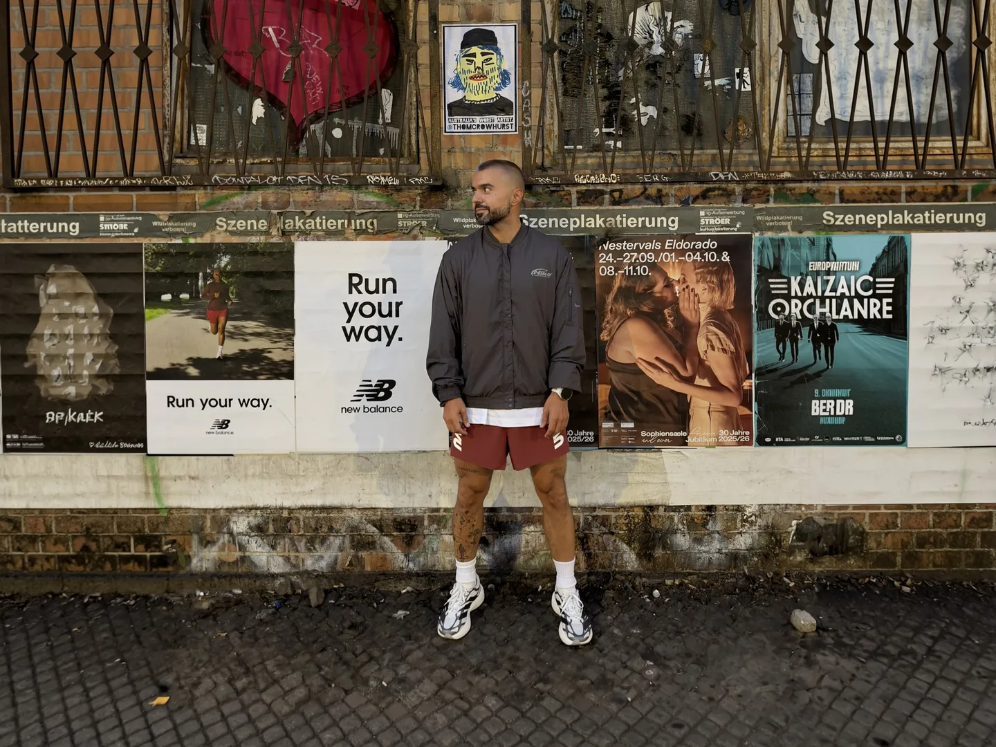 Serkan in Sportkleidung vor einer mit Plakaten beklebten Backsteinwand.
