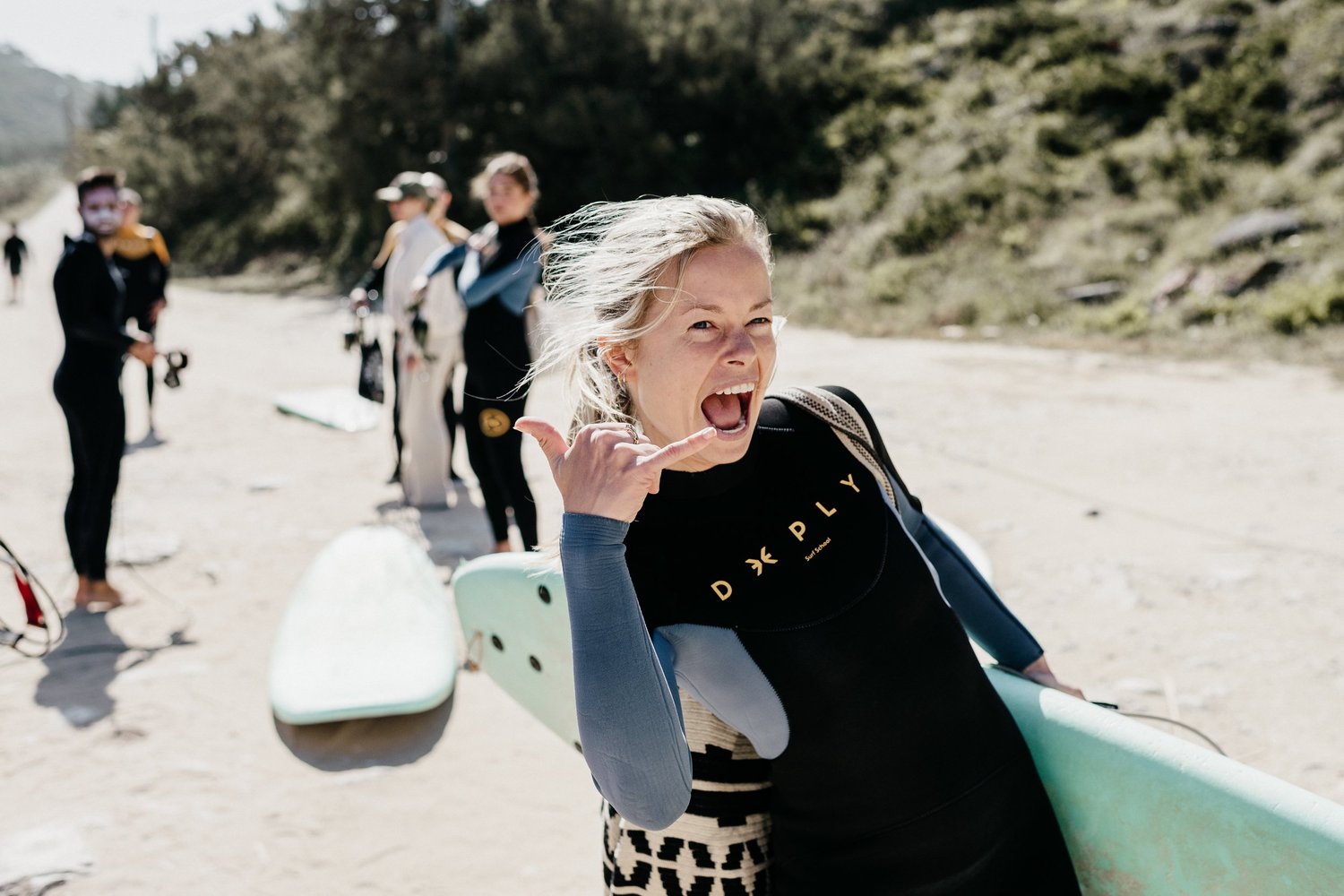 A woman in a wetsuit pulling a shaka sign with a surfboard under her arm