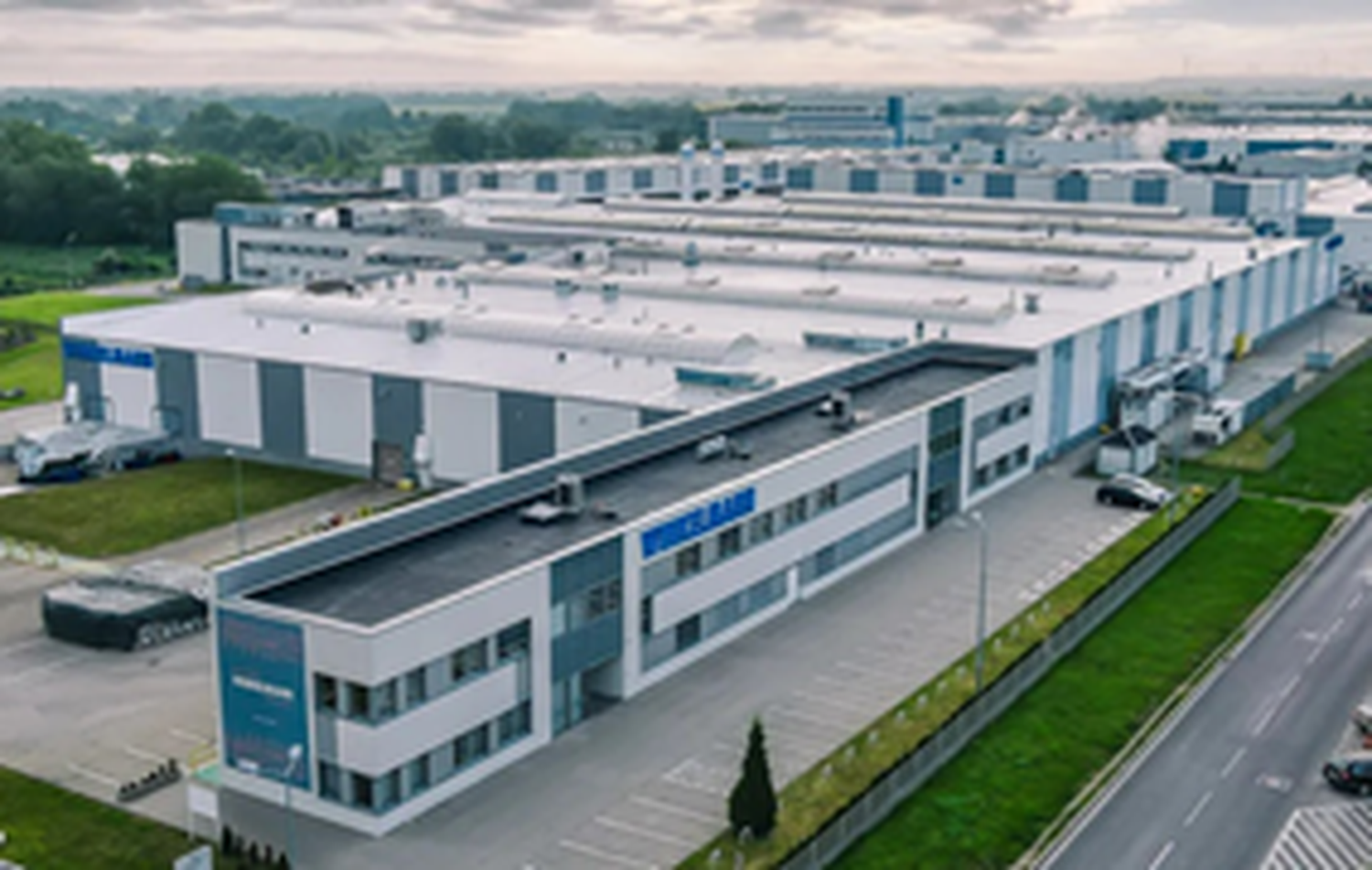 Luftaufnahme eines großen Industriekomplexes mit mehreren weißen und grauen Gebäuden, umgeben von Grünflächen und angrenzend an eine Straße.